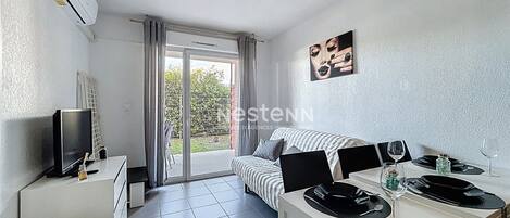 Een moderne woonkamer met een bank, een televisie, een eettafel gedekt voor twee personen en een glazen schuifdeur naar een balkon.
