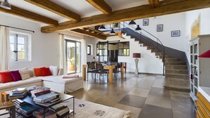 Ein großzügiger Wohnbereich mit hoher Decke, freiliegenden Holzbalken, einer Treppe, einem Essbereich und einer Couch mit Kissen.