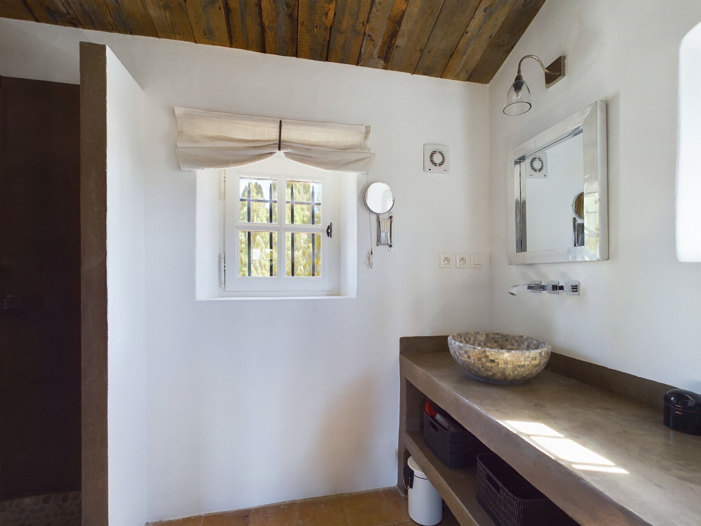 A bathroom with a window, a mirror, a sink with a mosaic bowl, and a wooden ceiling.