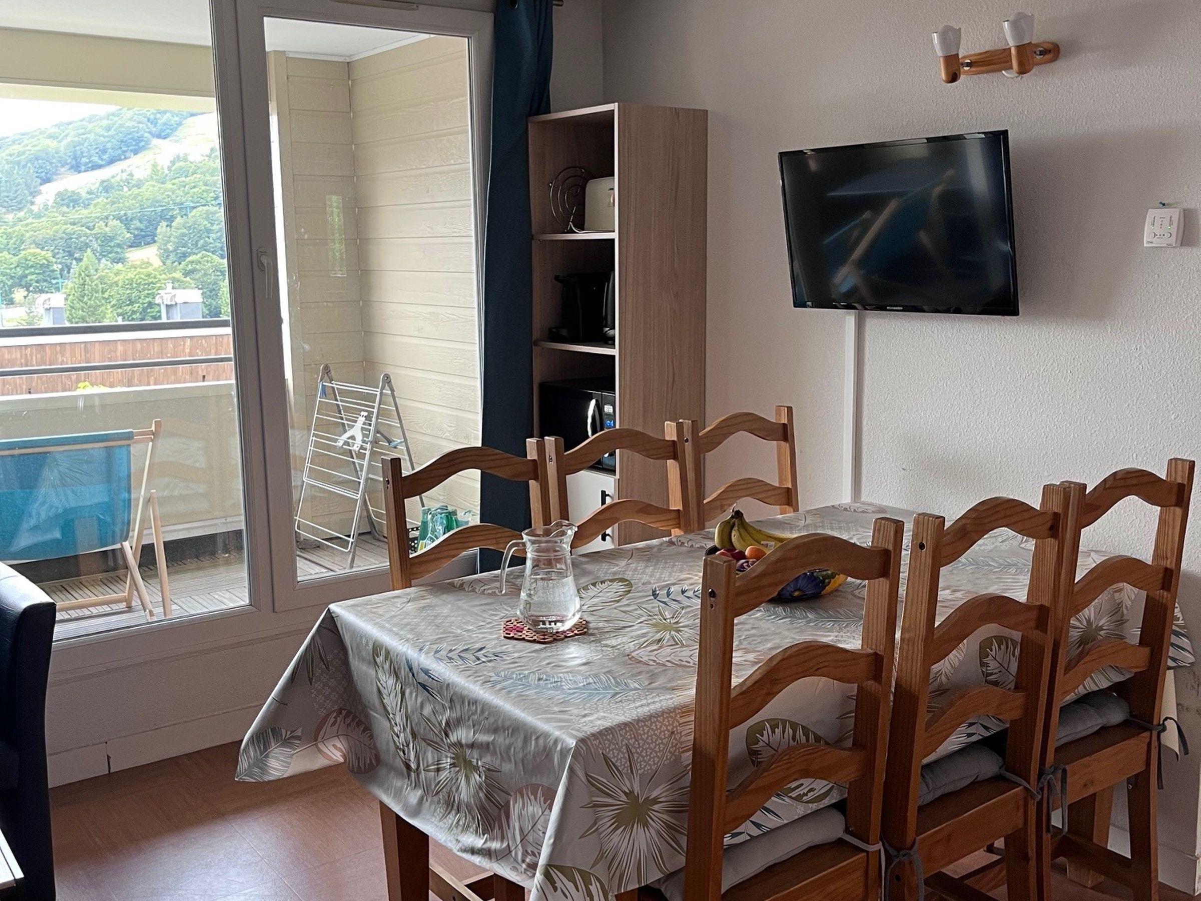 A dining area with a table set for four, a television mounted on the wall, and a view of a balcony with a pool.