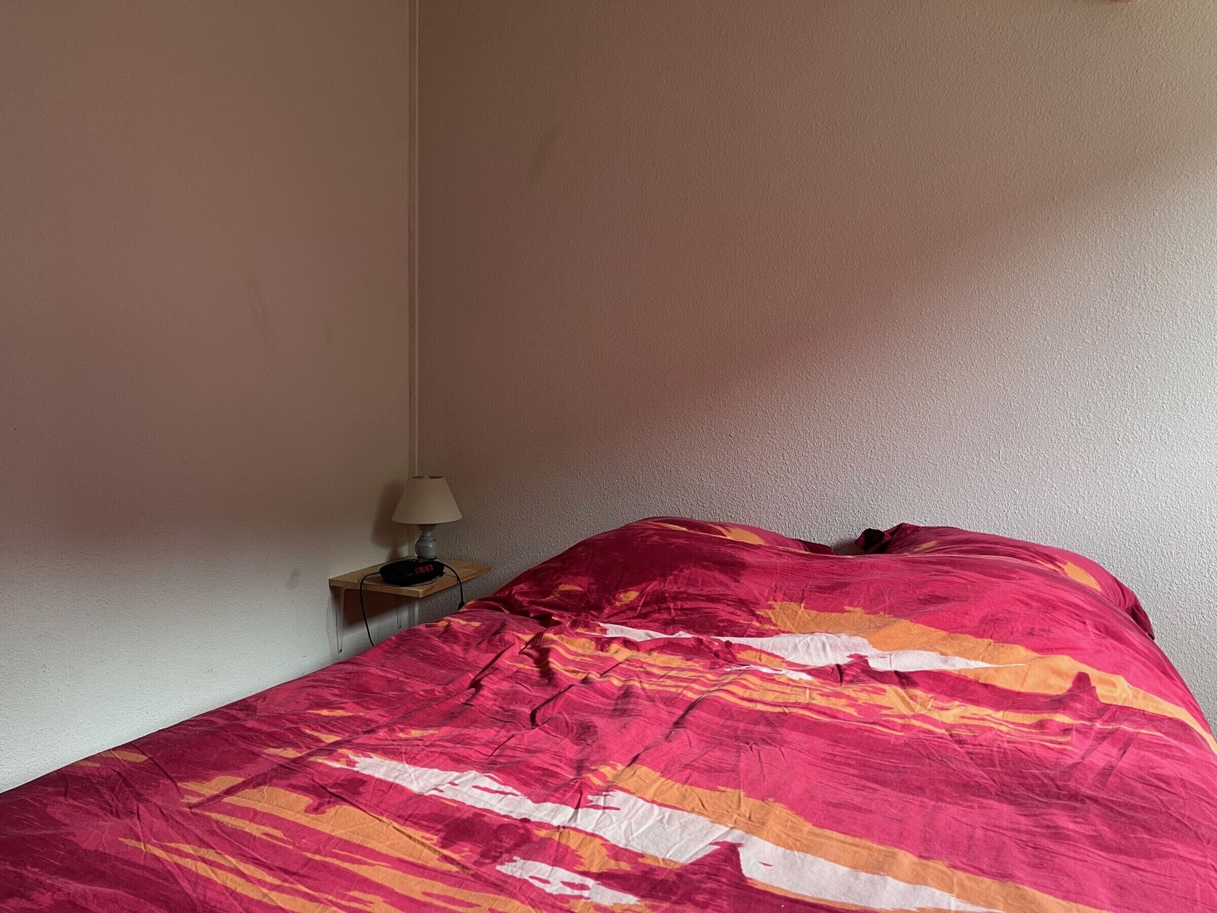 Una cama con una colcha estampada en rojo y naranja, una mesita de noche con una lámpara y una pared lisa.