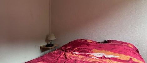 Una cama con una colcha estampada en rojo y naranja, una mesita de noche con una lámpara y una pared lisa.