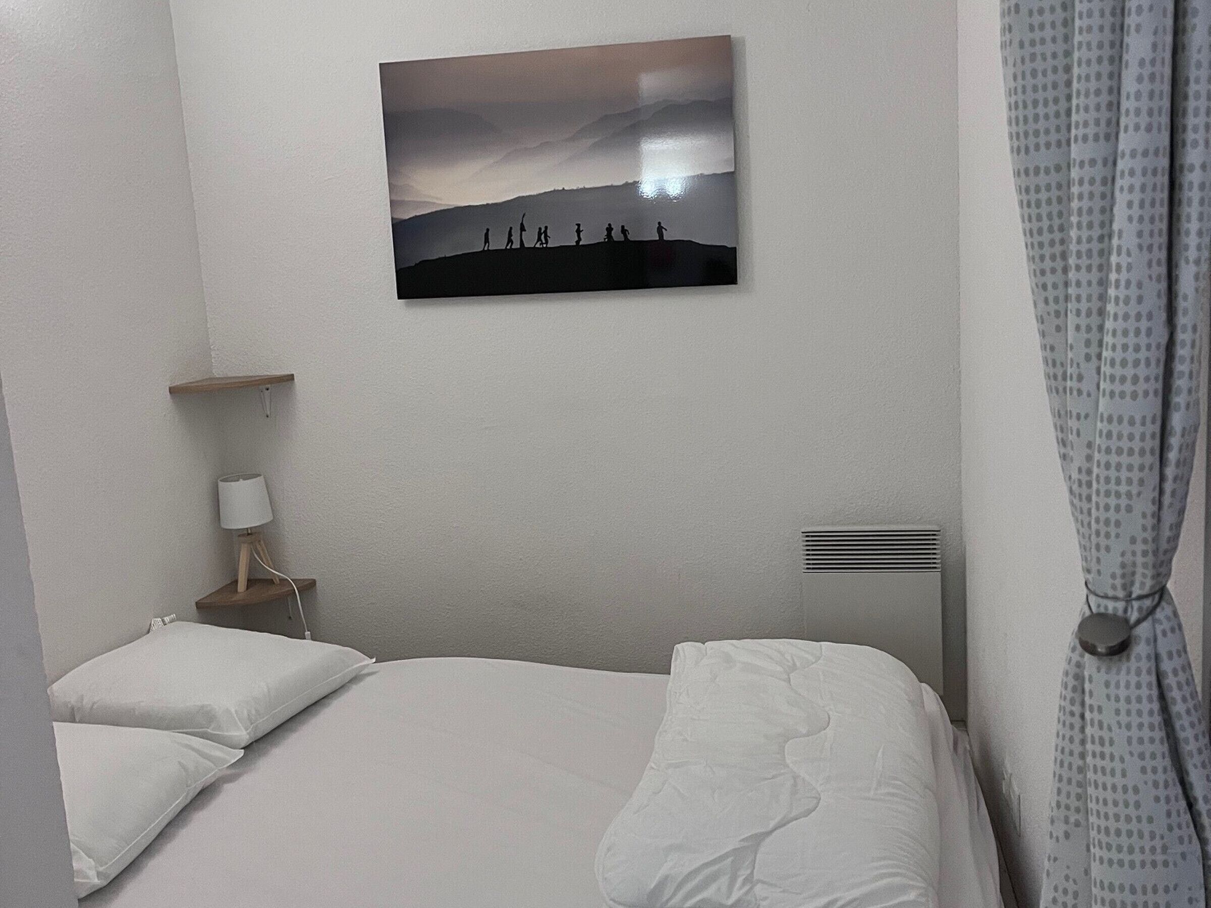 A hotel room with a bed, a bedside table with a lamp, a wall-mounted picture, and a curtain.