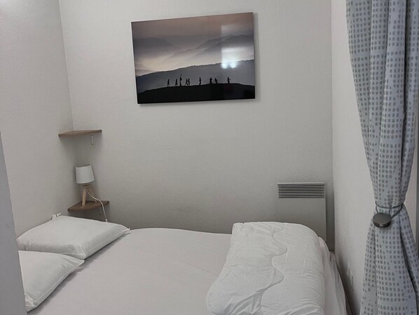 A hotel room with a bed, a bedside table with a lamp, a wall-mounted picture, and a curtain. - Spacious apartment for 6 with terrace and private parking (Besse-et-Saint-Anastaise-Super Besse)