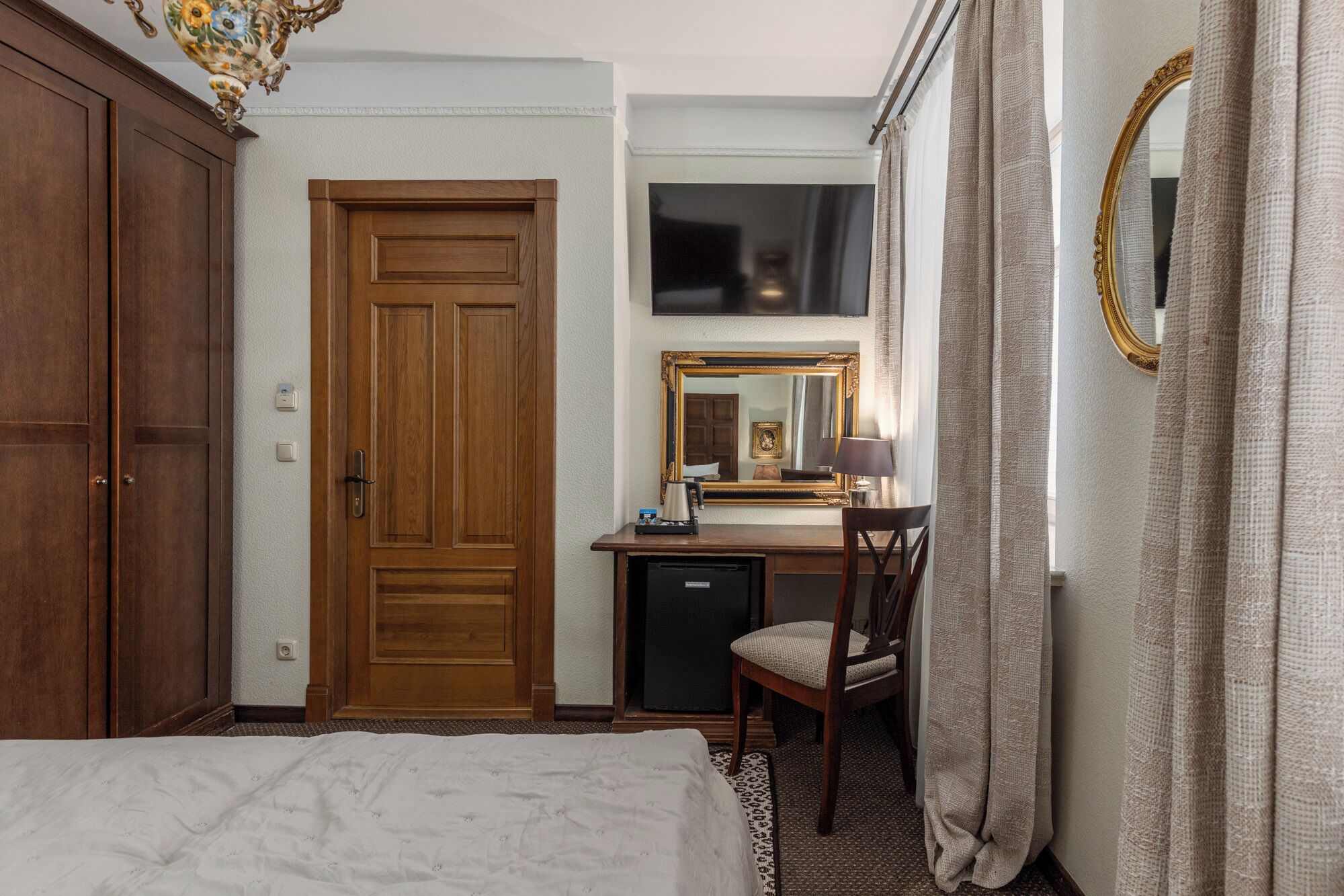 A hotel room with a wooden wardrobe, a desk with a chair, a bed, a television, and a large mirror.