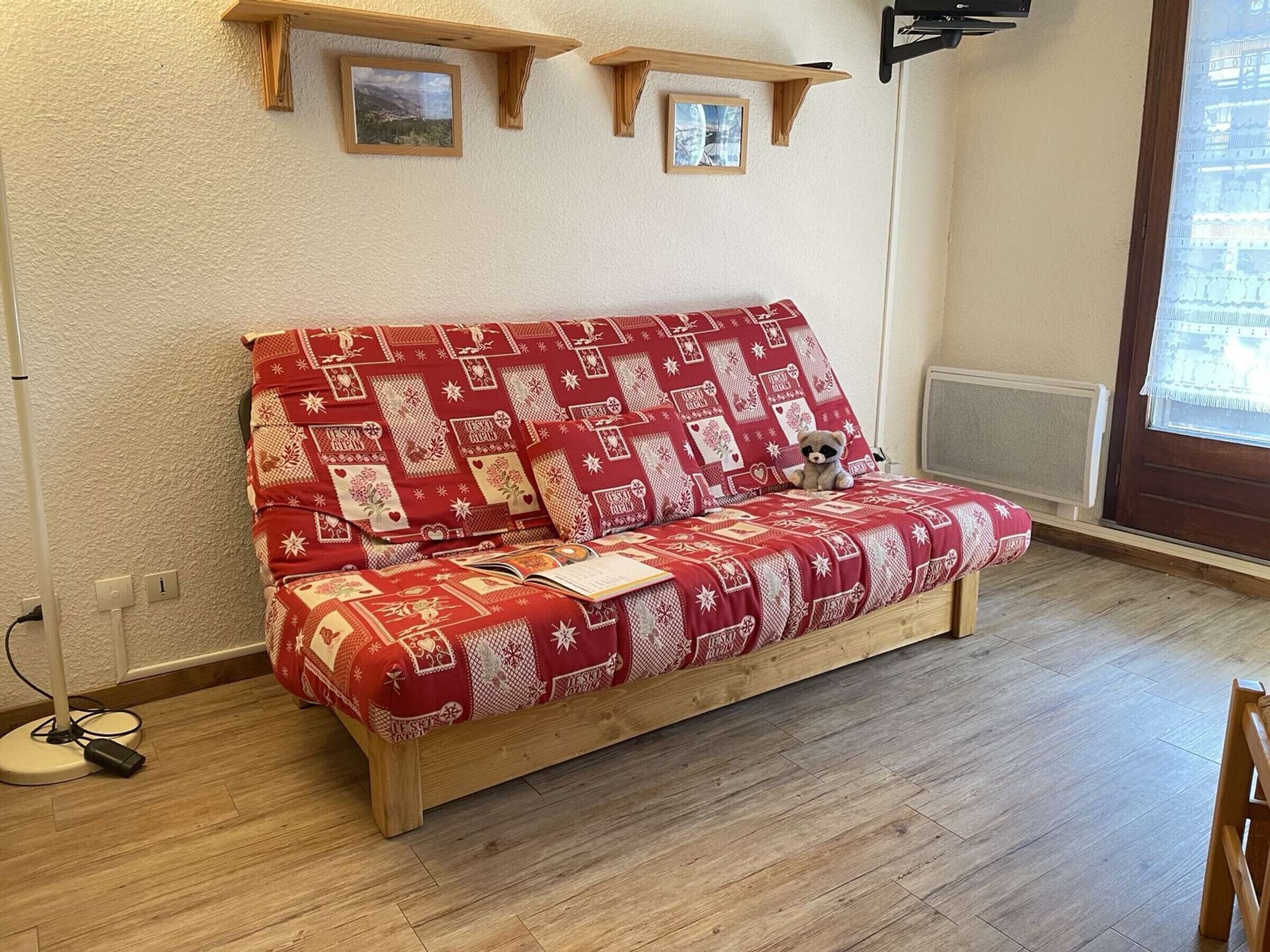 Un canapé en bois recouvert d’un tissu à motifs rouges, un petit ours en peluche posé dessus et un livre à côté. Au-dessus du canapé, on trouve deux étagères en bois, chacune portant un tableau encadré.