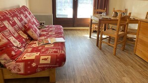 A cozy room with a red patterned sofa, a wooden dining table with chairs, and a view of buildings through a large window. - Mountain studio 4 pers, South, Ski-in/out (Risoul)