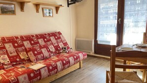 A cozy room with a wooden sofa covered in a patterned blanket, a small table with a chair, and a window with a view of a building. - Mountain studio 4 pers, South, Ski-in/out (Risoul)