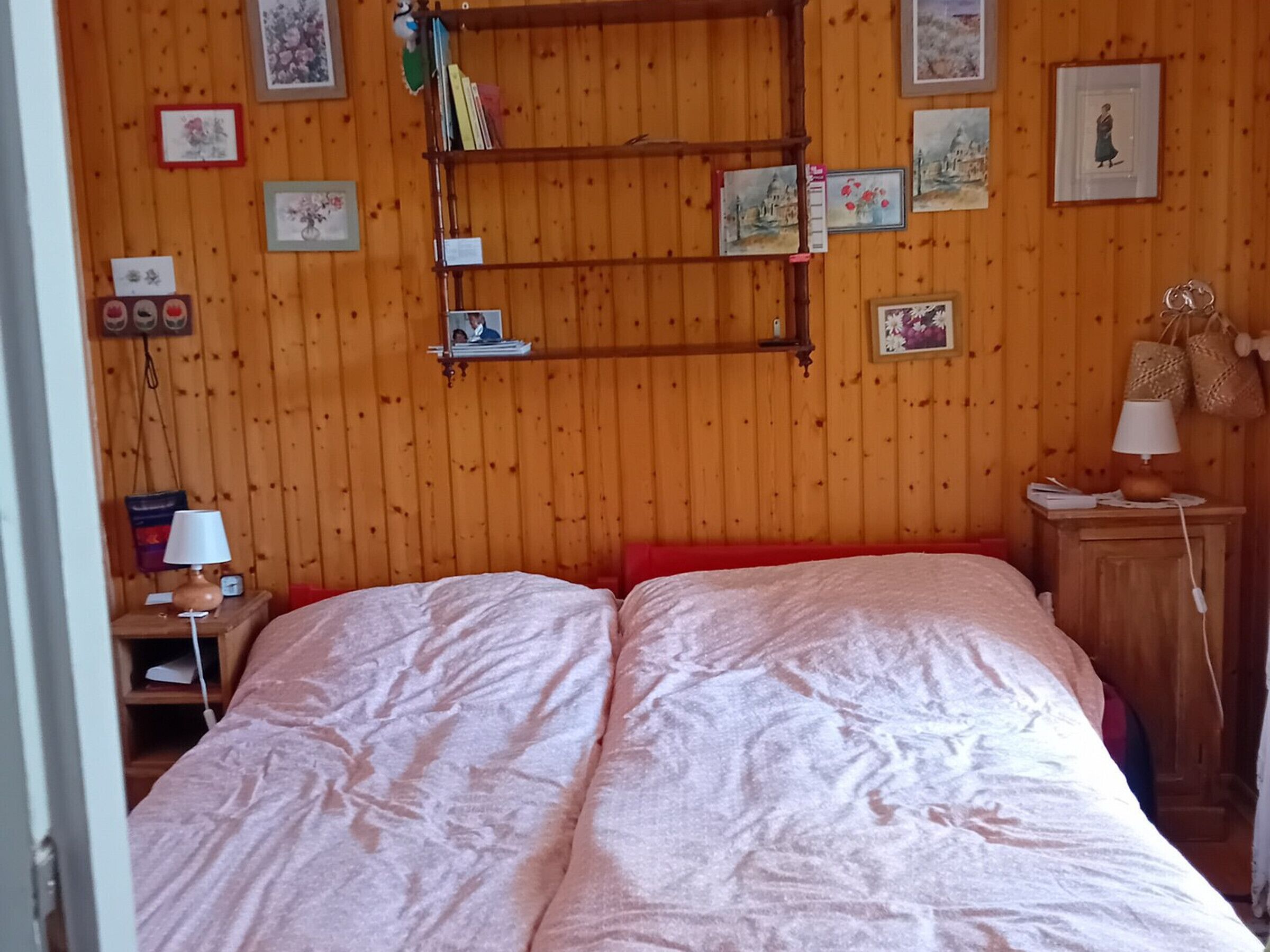 Una stanza con pareti in legno, un letto con testiera rossa, un comodino con una lampada e mensole con quadri incorniciati e libri.