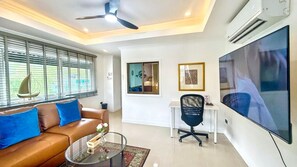 A modern living room with a brown leather sofa, a glass coffee table, a black office chair, a large flat-screen TV, and a ceiling fan.