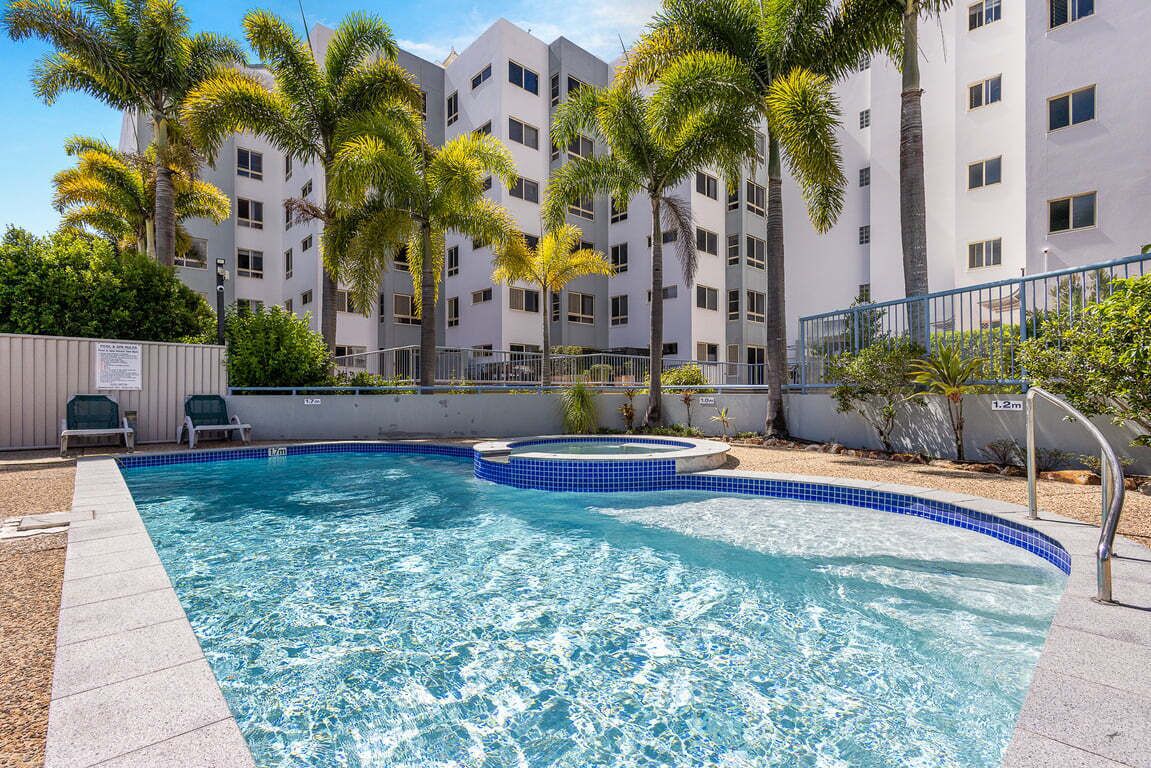 A swimming pool with a blue tiled edge, surrounded by palm trees and a multi-story building in the background.