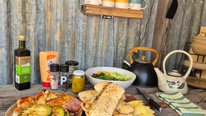 Ambiente rústico de cocina con una mesa de madera que exhibe diversos alimentos, incluyendo pollo a la parrilla, verduras y pan. Hay condimentos, una botella de aceite de oliva y una tetera sobre la mesa.