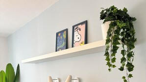 A bedroom with a blue headboard, two framed artworks, a hanging plant, bedside lamps, a bedside table with a clock and a jar, and a large green plant in a pot. - JUPAS° | Saartal Suite - Smart & Netflix (Konz)