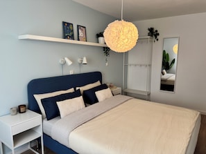 A modern bedroom with a large bed, a bedside table, a nightstand, a hanging light fixture, a shelf with artwork, and a mirror. - JUPAS° Saartal Suite – Quiet, Playground & Parking (Konz)