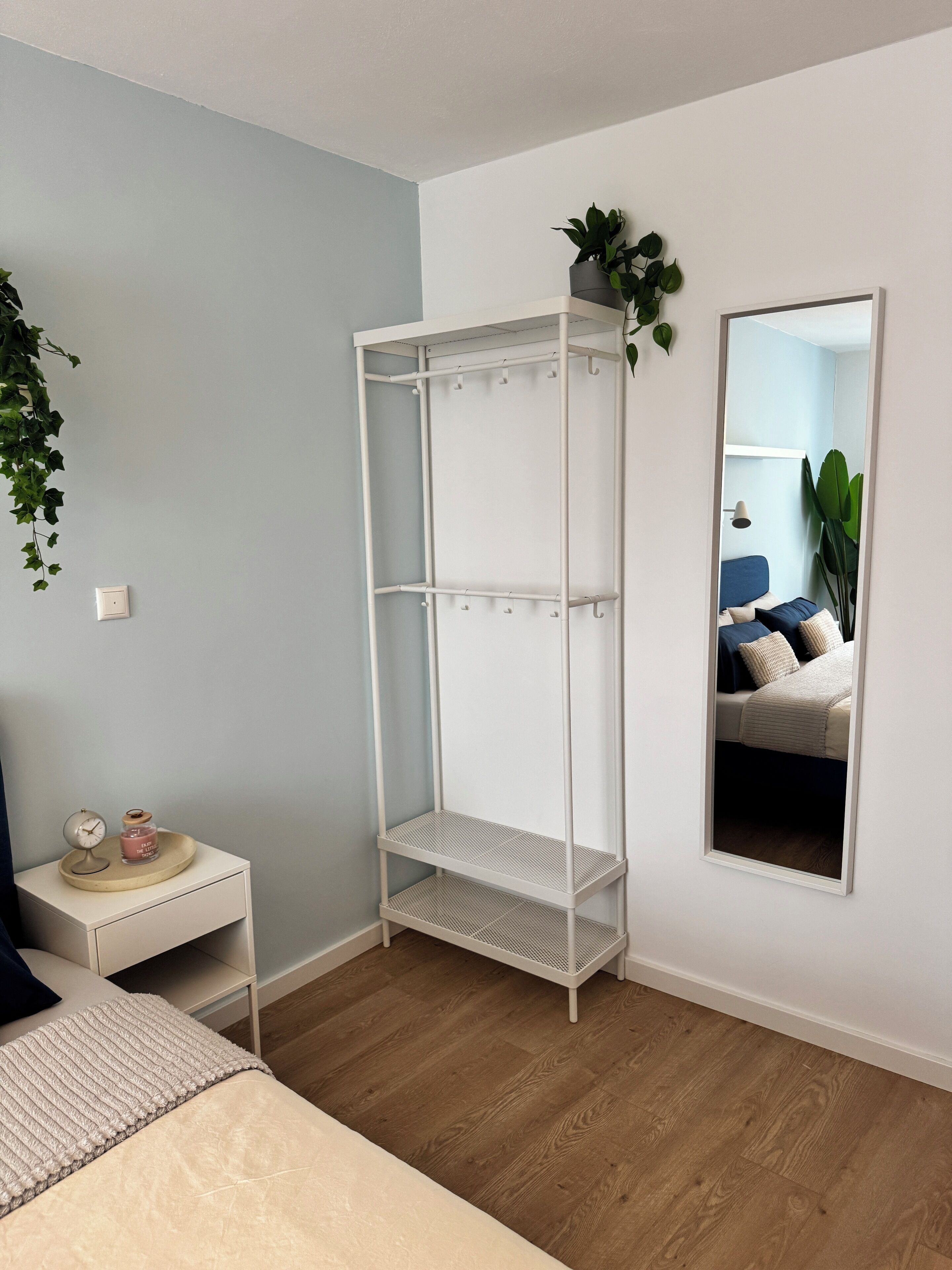 Une chambre à coucher avec un lit, une armoire en métal blanche, une table de chevet avec une horloge et une petite plante, et un miroir qui reflète la pièce.