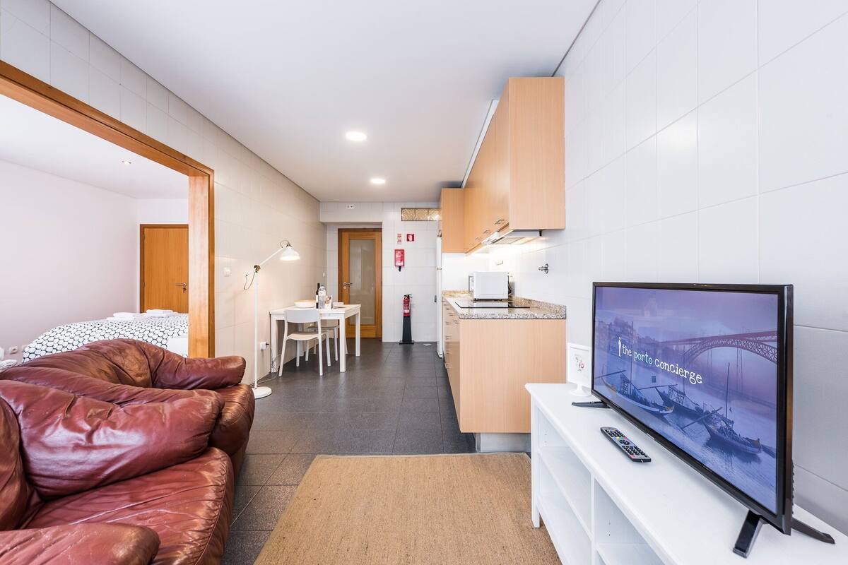 A modern hotel room with a leather sofa, a TV displaying a cityscape, a kitchenette, and a dining area.