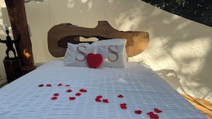 Ein Bett mit weißer Bettdecke, zwei Kissen mit Herz-Applikation und Rosenblüten, die auf dem Bett verteilt sind.