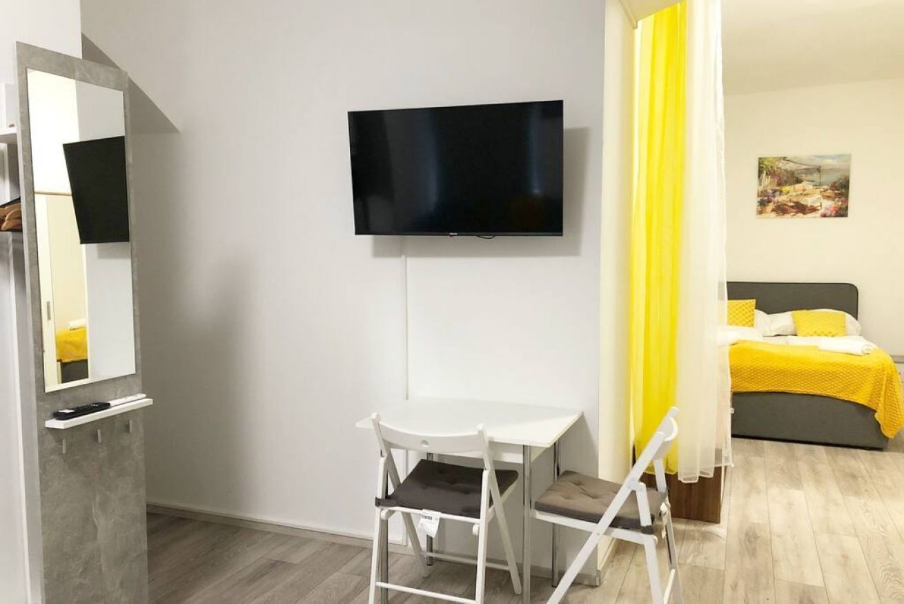 Una habitación con una televisión montada en la pared, un escritorio blanco con una silla, una cama con colcha amarilla y un cuadro en la pared.