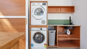 A compact laundry room with two front-loading washing machines, a sink, and a small trash bin. - Drift Studio - Cosy Fremantle Hideaway (Beaconsfield)