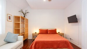 A bedroom with a bed, a wooden wardrobe, a TV mounted on the wall, and a white sofa with a blue pillow. - Drift Studio - Cosy Fremantle Hideaway (Beaconsfield)