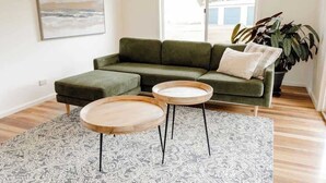 A living room with a green sofa, two round wooden side tables, a patterned rug, and a large window with a view.
