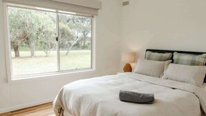 A bedroom with a large window, a bed with white bedding, a lamp, and a view of trees outside.