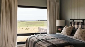 Quarto com uma grande janela, cama com colcha xadrez, mesa de cabeceira com abajur e vista para uma paisagem gramada.
