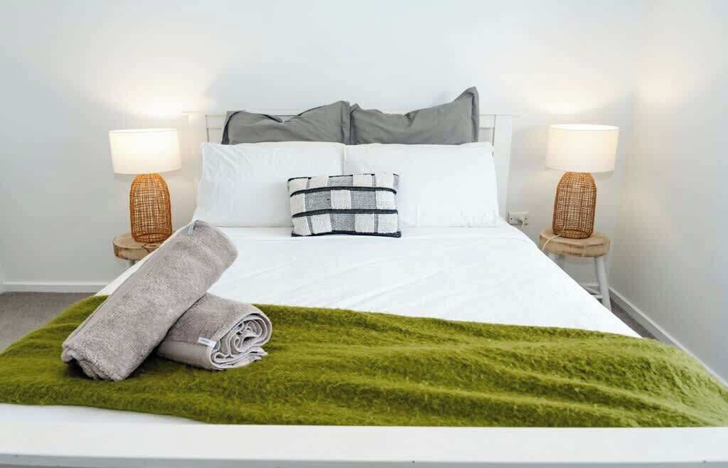 A neatly made bed with white bedding, two pillows, a green throw blanket, and two bedside lamps on matching nightstands.