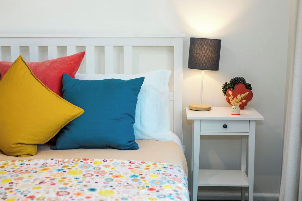 Una cama con almohadas coloridas, una colcha estampada, una mesita de noche con una lámpara y un pequeño objeto decorativo.