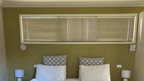 A double bed with a grey headboard, two bedside tables with lamps, a window with blinds, and a wall decoration.