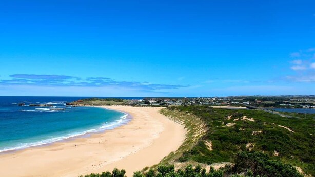 A coastal landscape with a sandy beach, a clear blue sky, and a distant view of a town.