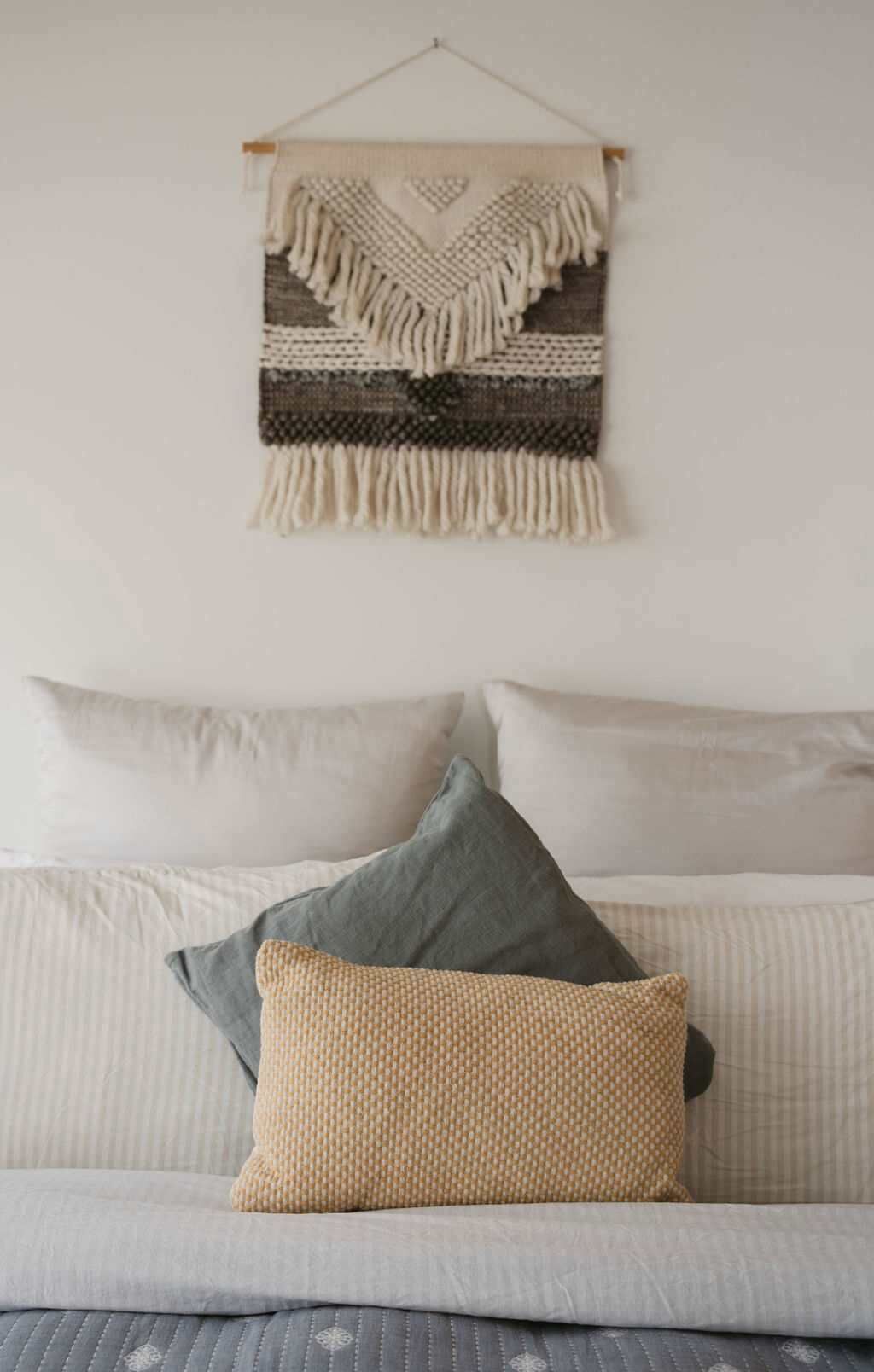 A bed with a grey and white striped bedspread, a beige woven throw pillow, and a grey pillow. Above the bed, there is a wall hanging with a geometric pattern and fringe.