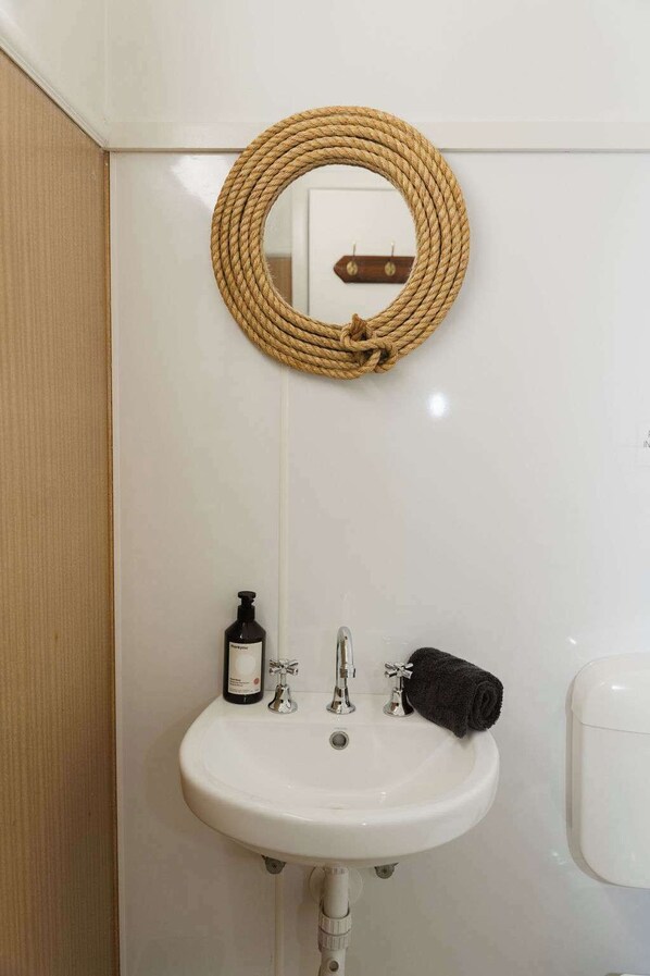 A bathroom with a white sink, a mirror, and a wooden rope decoration. - Neptunes Guest House (Port Campbell)