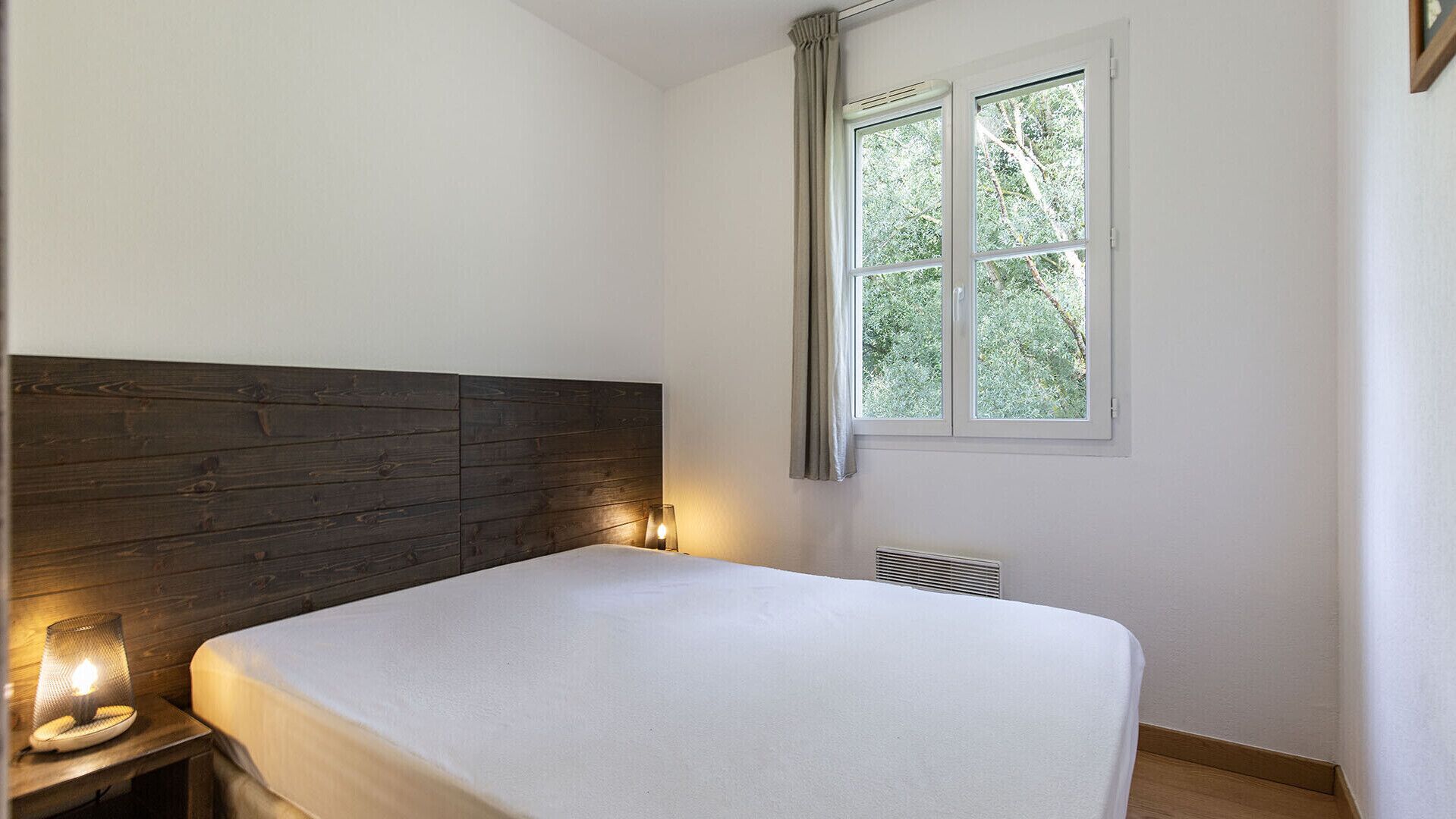 A bedroom with a wooden headboard, a bed with a white mattress, a bedside table with a lamp, and a window with curtains.