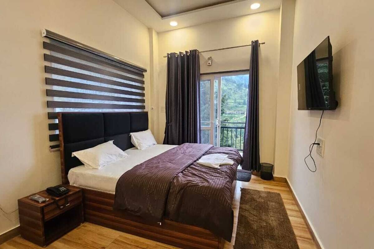 Um quarto com cama, mesa de cabeceira, televisão na parede e janela com cortinas.