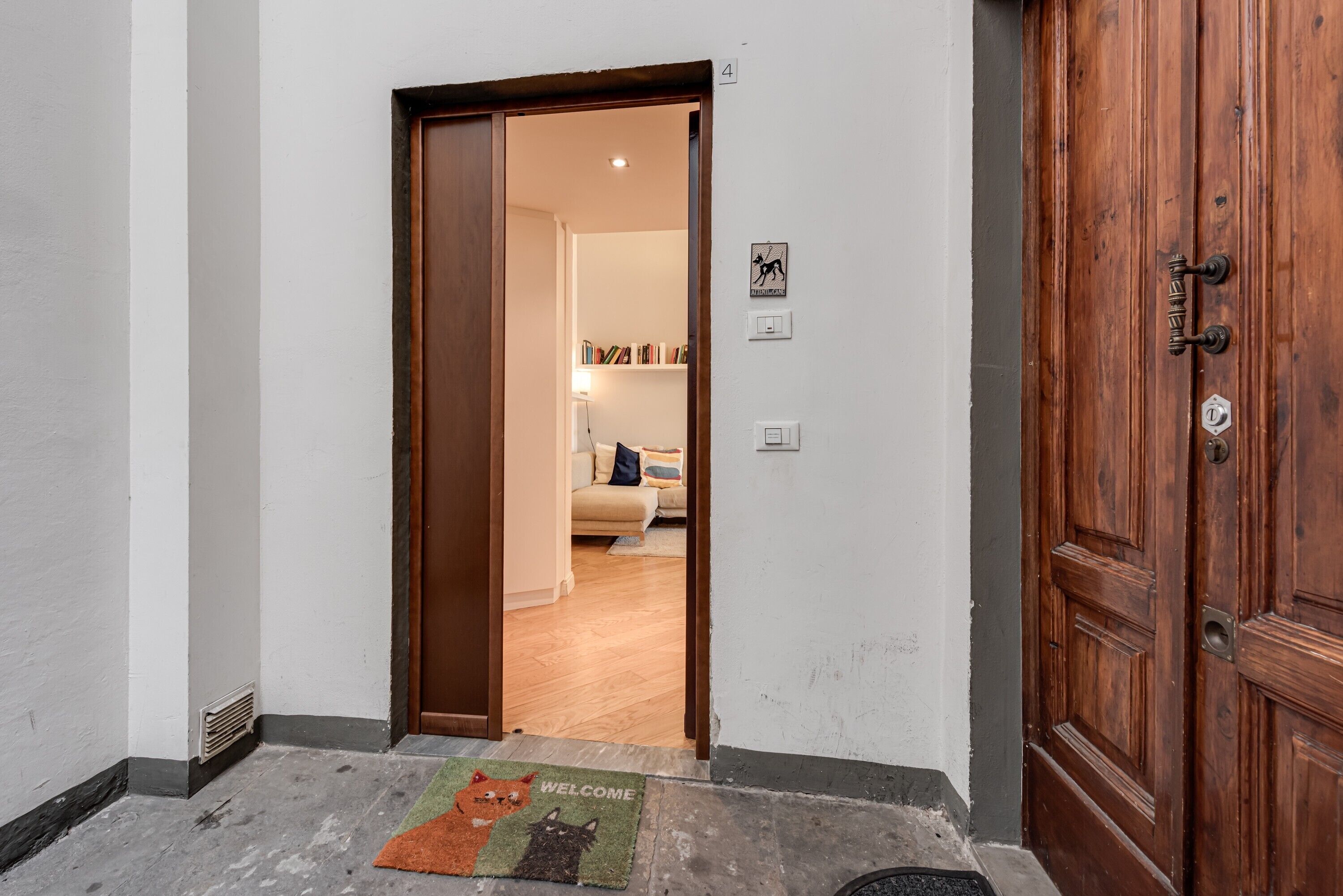 A hallway with a wooden door, a welcome mat, and a view into a living room with a sofa and bookshelves.