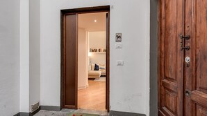 A hallway with a wooden door, a welcome mat, and a view into a living room with a sofa and bookshelves.