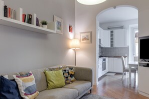 Uma sala de estar moderna com sofá, luminária, estante de livros e área de cozinha visível por um arco.