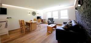 Un salon moderne avec un plancher en bois, un canapé noir, une table à manger avec des chaises, un téléviseur et un mur en pierre.