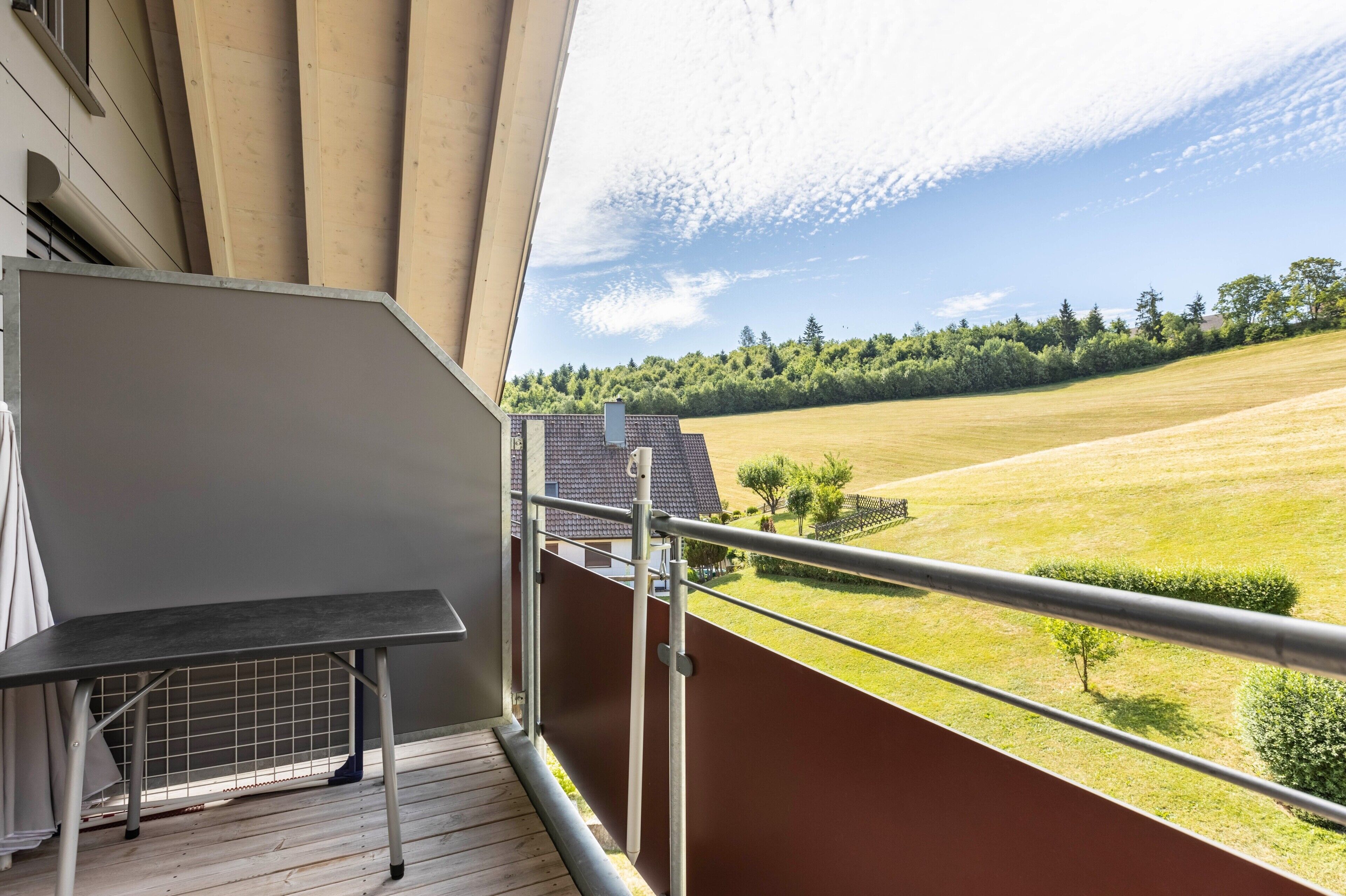 A balcony with a metal table, a view of a green field, and a building with a sloped roof.