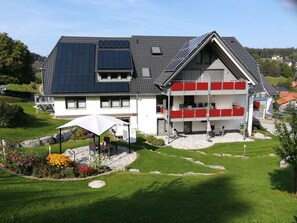 A modern two-story house with solar panels, a patio with outdoor furniture, and a well-maintained garden.