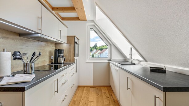 Une cuisine étroite avec un plancher en bois, des armoires blanches, un comptoir noir et une fenêtre donnant sur des maisons.