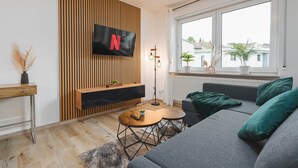 Ein modernes Wohnzimmer mit Wand-Fernseher, Holzwandverkleidung und einem großen Fenster mit Blick auf Gebäude.
