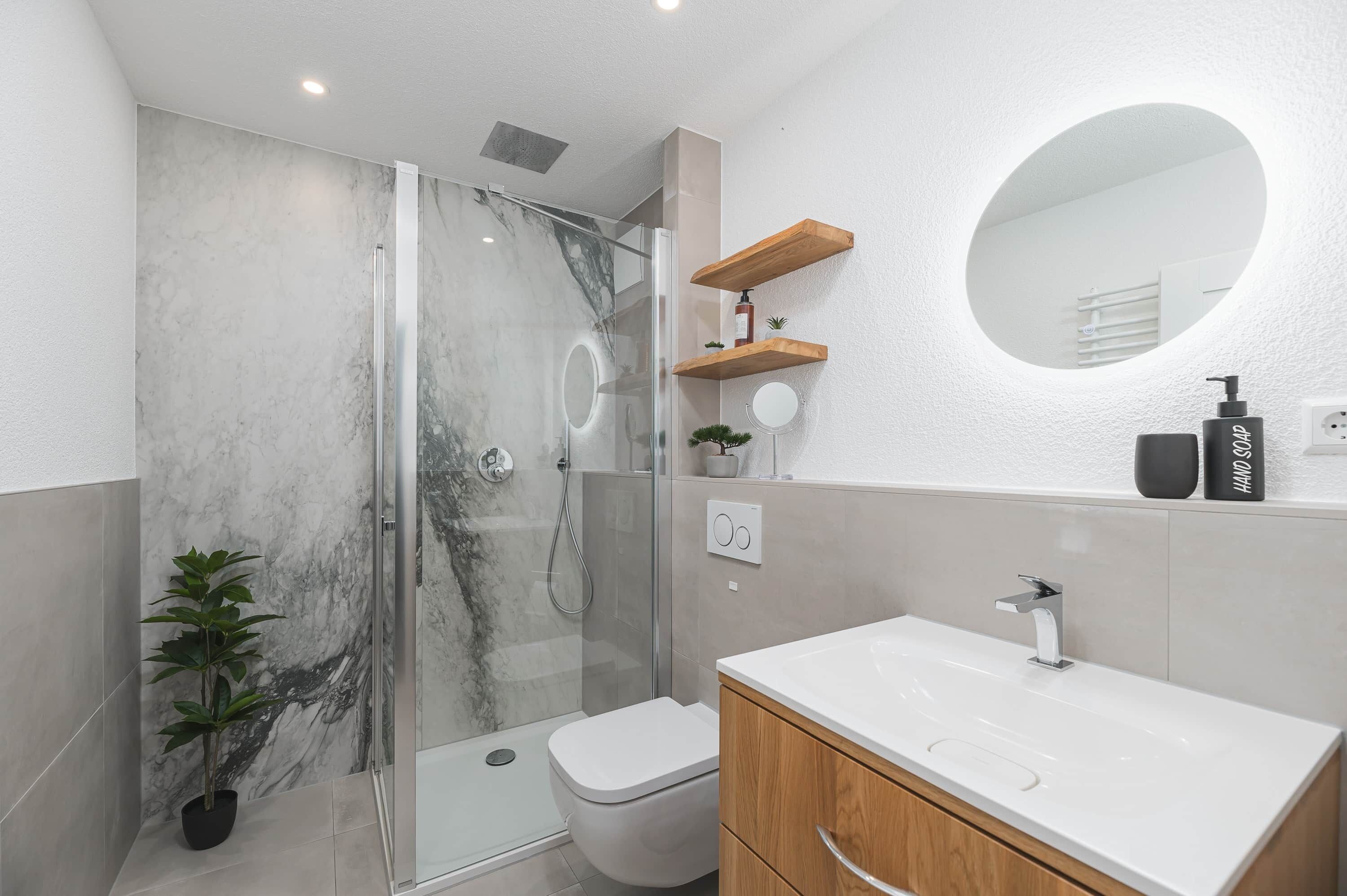 A modern bathroom with a glass-enclosed shower, a toilet, a sink with a wooden cabinet, and a round mirror on the wall.