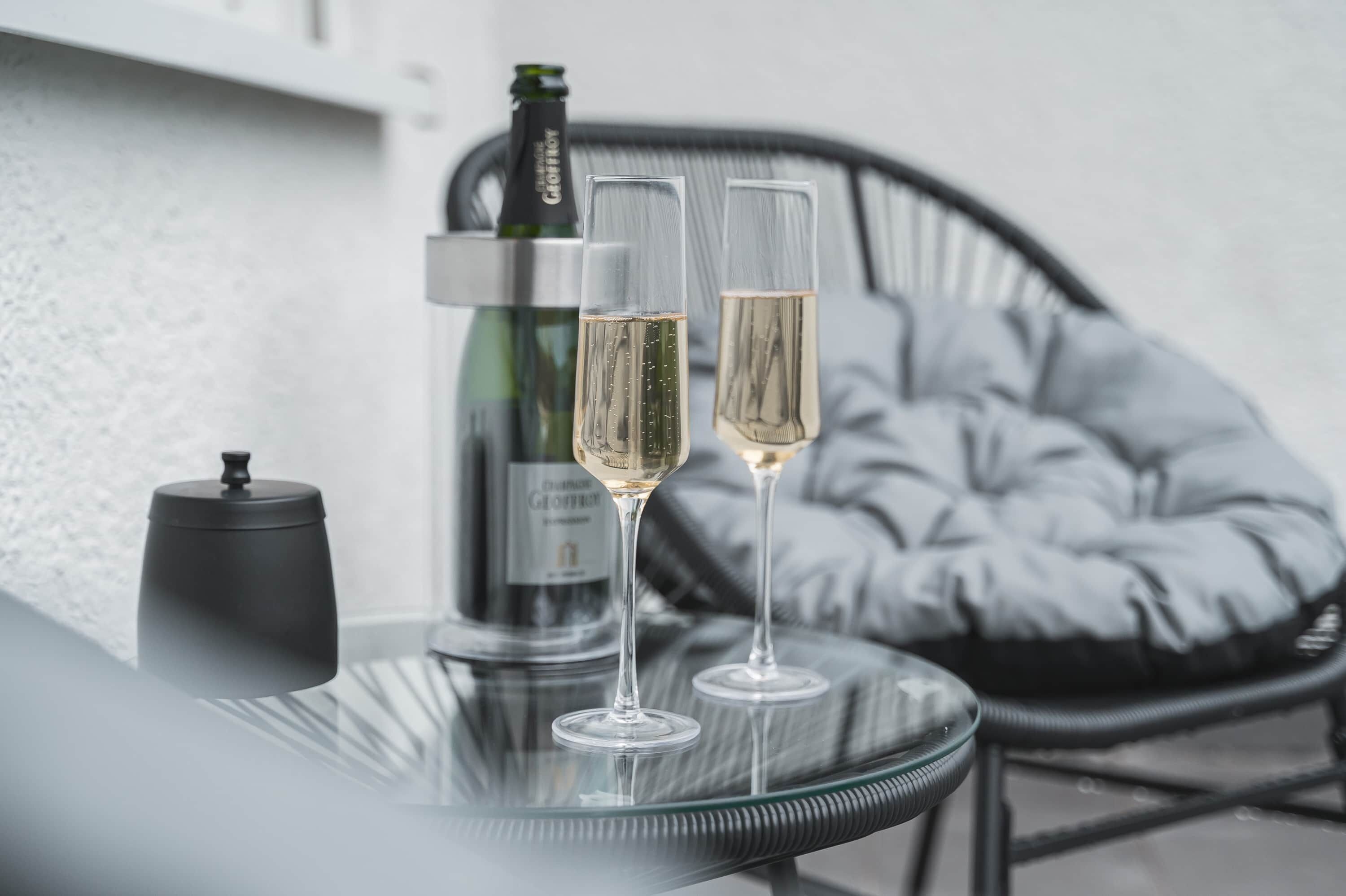 Ein Glastisch mit zwei Sektgläsern und einer Champagnerflasche. Auf dem Tisch steht ein schwarzer Behälter. Im Hintergrund ist ein schwarzer Stuhl mit Kissen zu sehen.