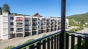 A multi-story residential building with balconies, a parking area, and a view of a hilly landscape.