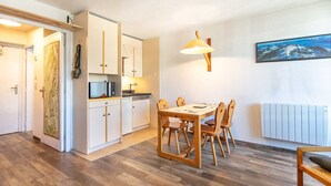 A compact kitchen with white cabinets, a dining table with four chairs, a hanging lamp, and a framed picture on the wall.