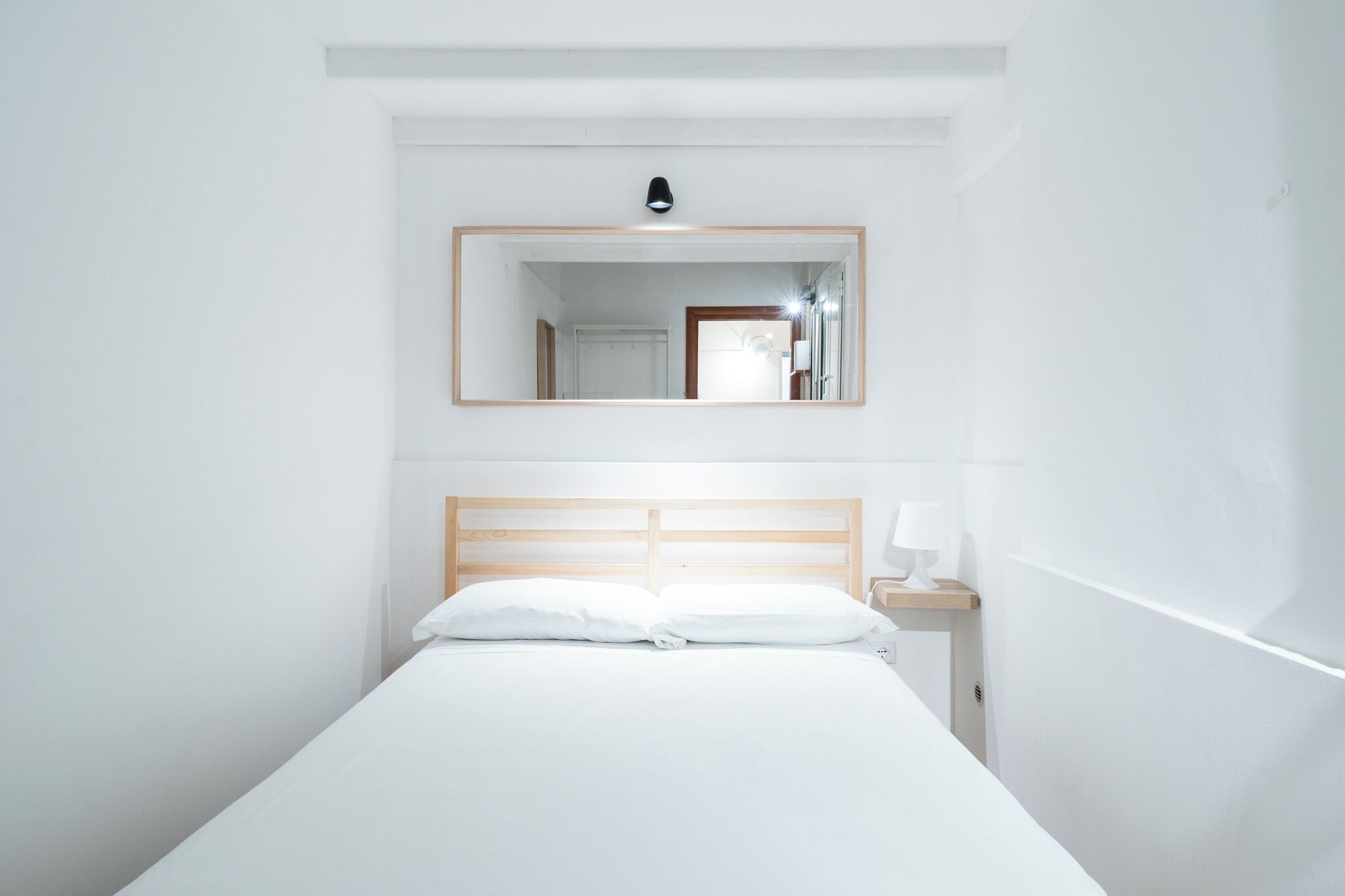 A minimalist hotel room with a single bed, a nightstand, a bedside lamp, and a mirror above the bed.