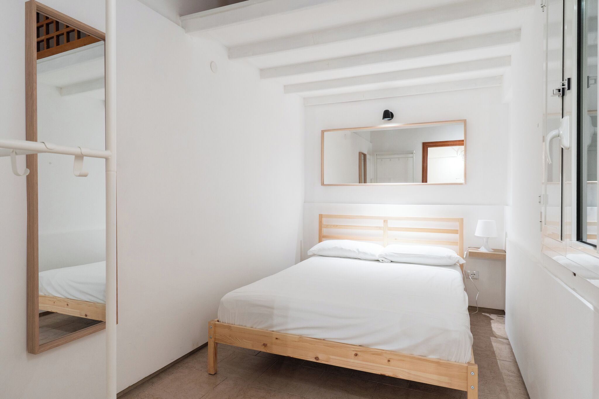 A bedroom with a wooden bed, a mirror, a bedside table with a lamp, and a window.