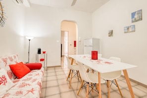 A modern living room with a white sofa, red cushions, a dining table with chairs, a lamp, and a refrigerator. - Apartment 'Corallo' with Private Terrace, Wi-Fi and Air Conditioning (Porto Cesareo)